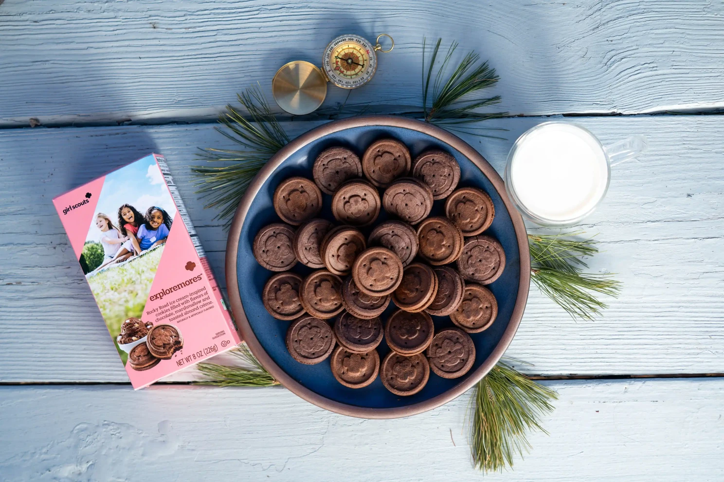 Girl Scout cookies in a bowl beside packaging, milk, and pine sprigs on a wooden tabletop.